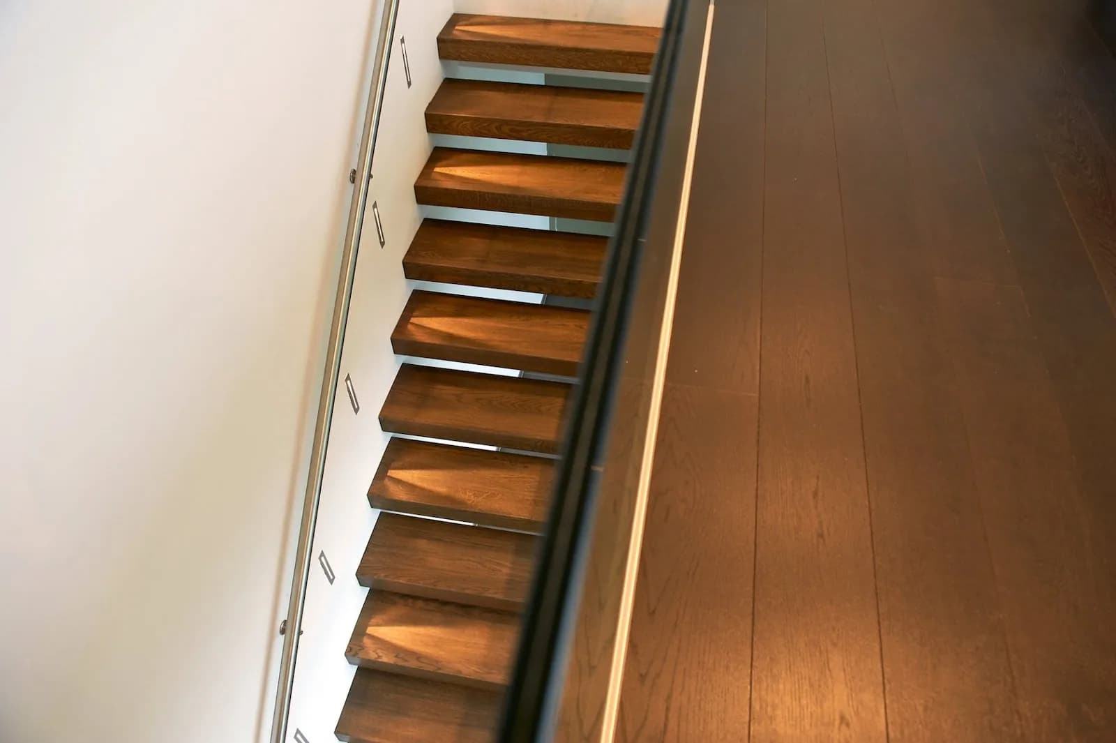 Marches en bois foncé vues d'en bas le long d'un mur blanc, garde-corps vitré, large plancher en bois foncé au premier plan