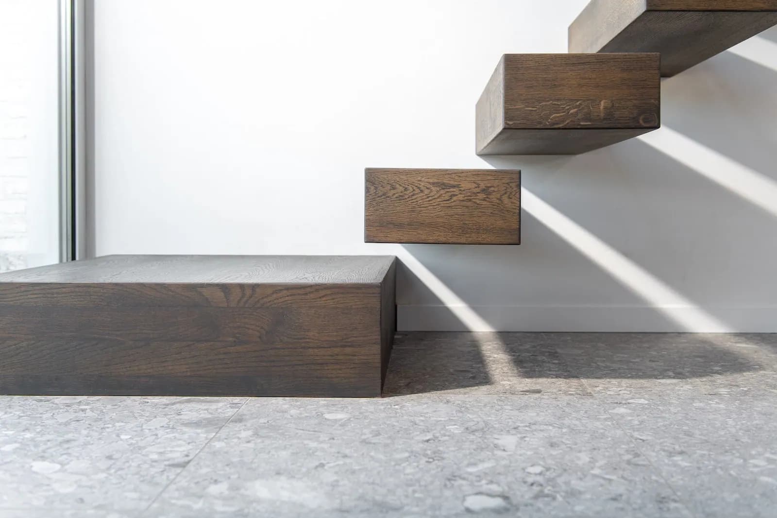 Première marche en bois foncé posée sur un sol en terrazzo gris, marches suivantes encastrées dans le mur blanc, lumière rasante