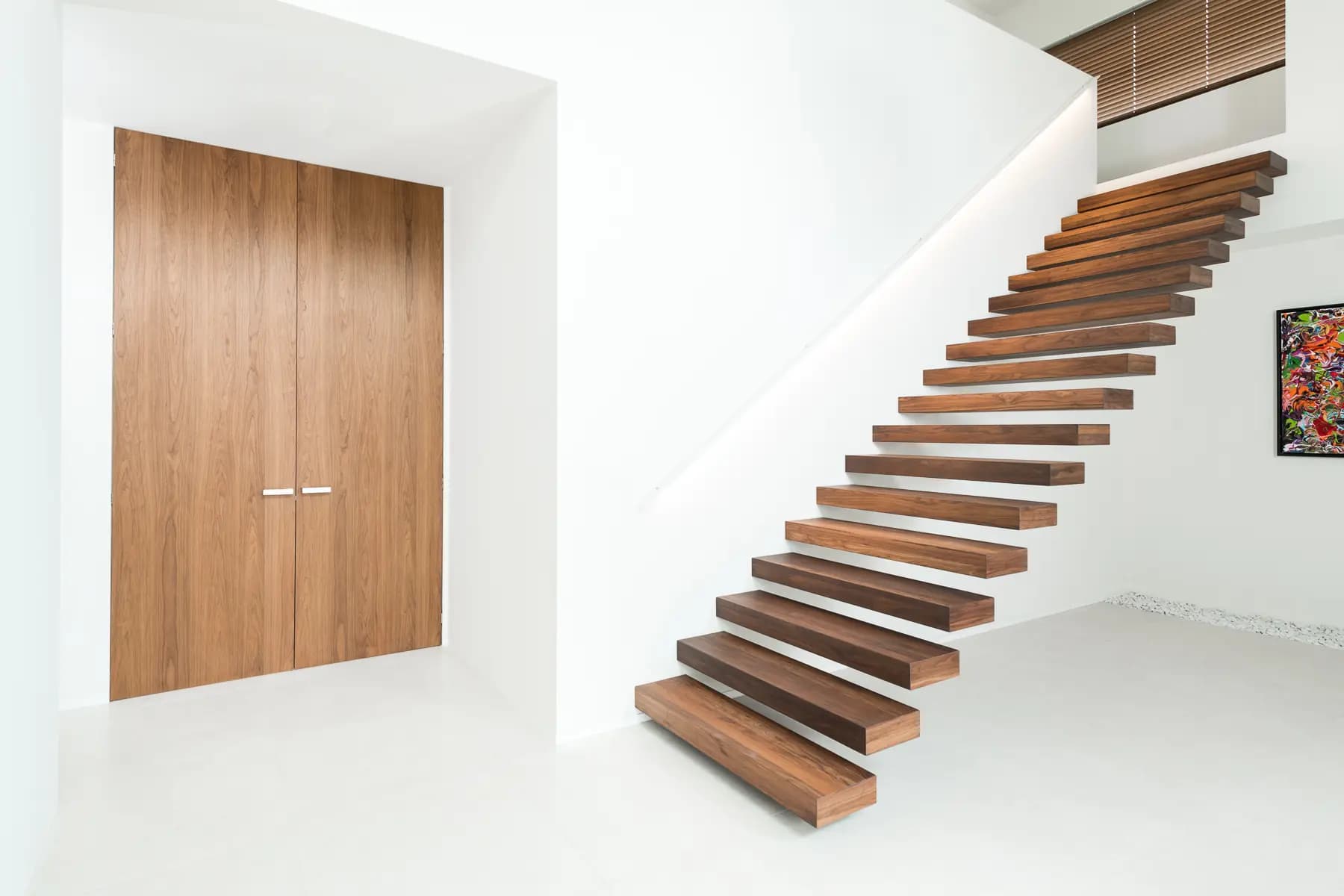 Escalier aux marches épaisses en bois foncé encastrées dans un mur blanc, face à une porte double pleine hauteur en bois