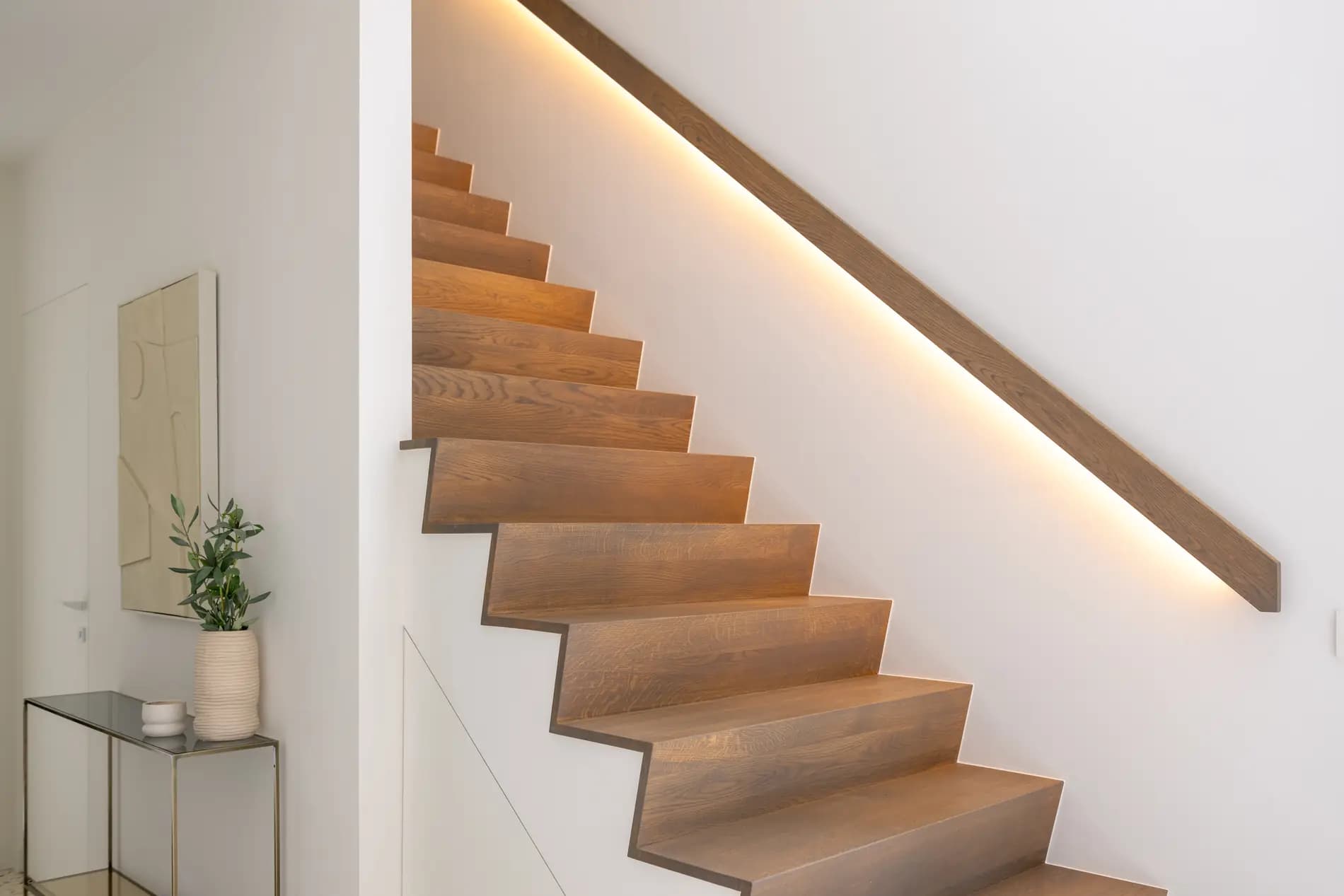 Escalier en bois clair montant le long d'un mur blanc, main courante plate en bois avec une ligne lumineuse dissimulée en dessous, console et petit vase au premier plan
