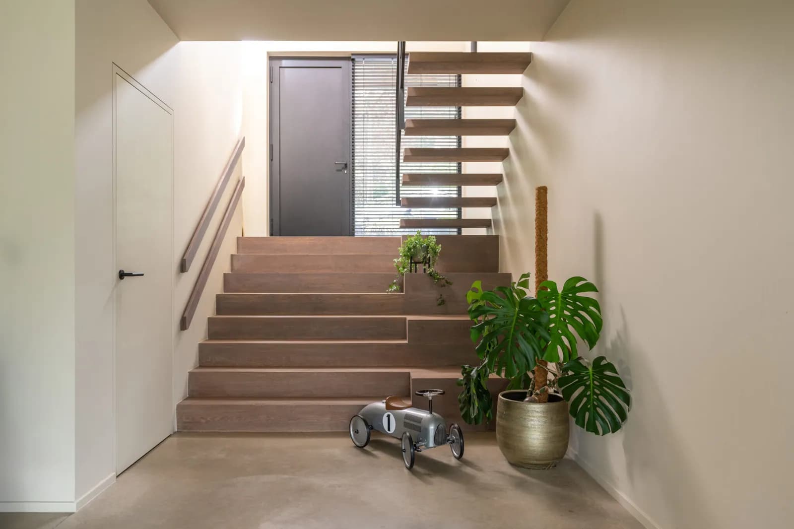 Escalier en bois menant à un palier, porte blanche sans encadrement apparent à gauche et porte grise en haut des marches, sol en béton ciré, grande plante verte