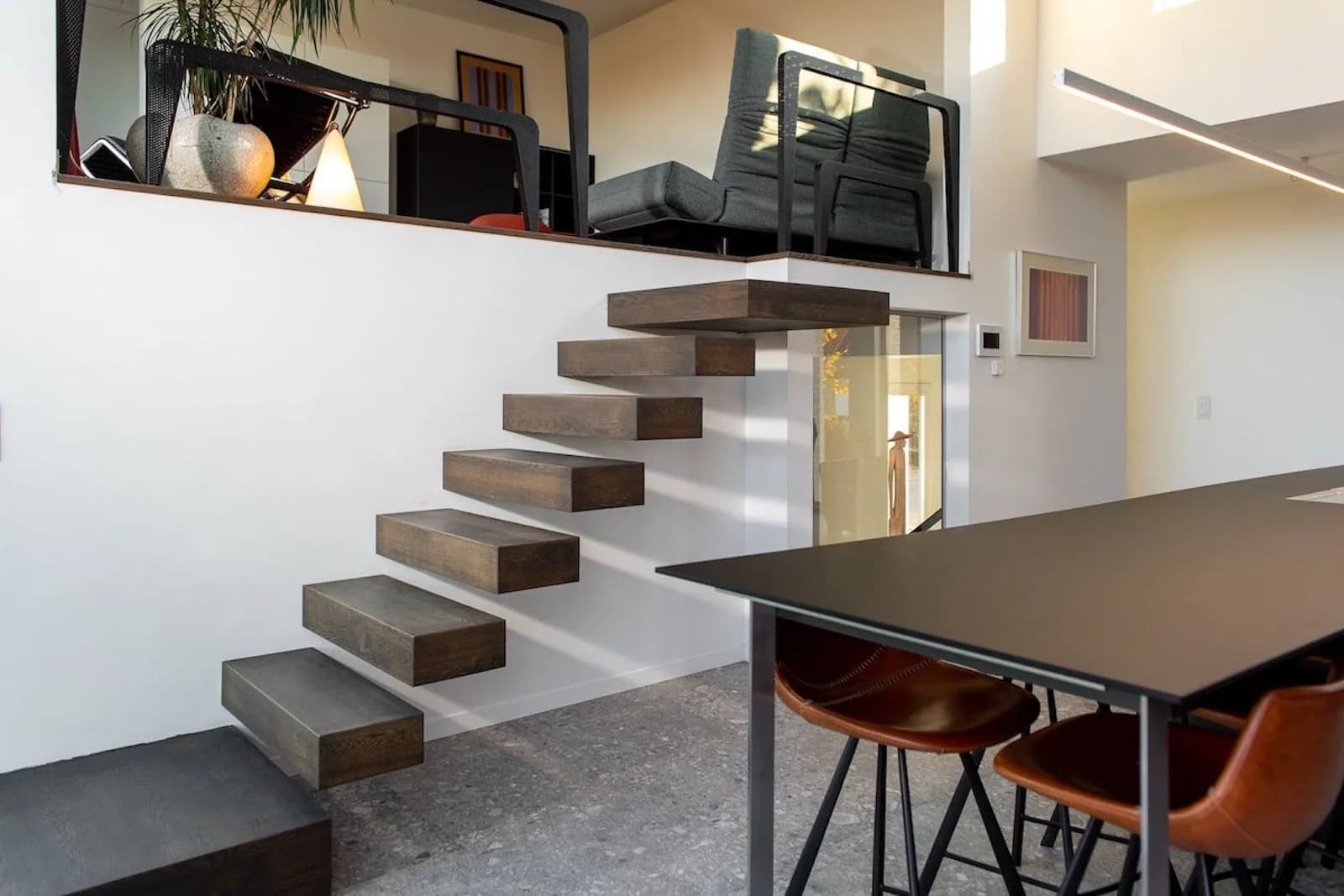 Escalier aux marches épaisses fixées dans un mur blanc, dans une pièce ouverte avec une longue table sombre et des tabourets en cuir, mezzanine à l'étage