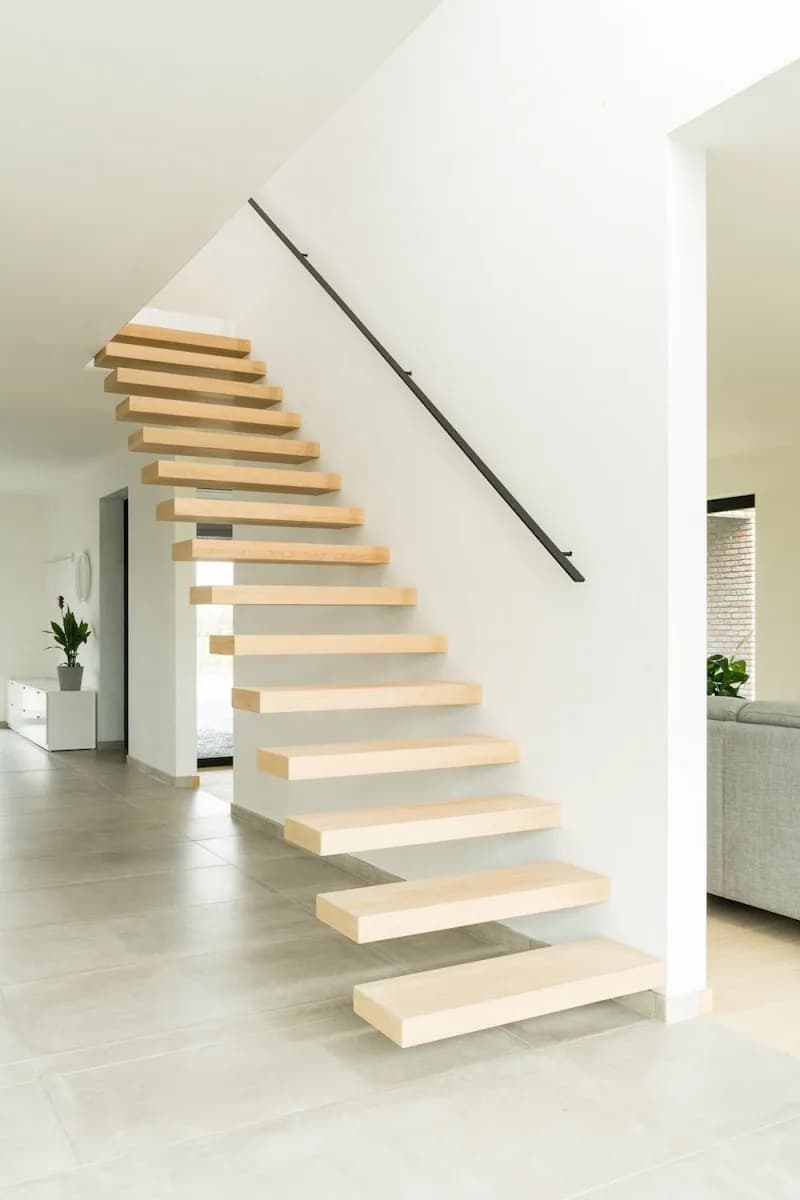 Escalier aux marches en bois clair fixées dans un mur blanc, fine main courante noire, sol carrelé clair et séjour visible au fond
