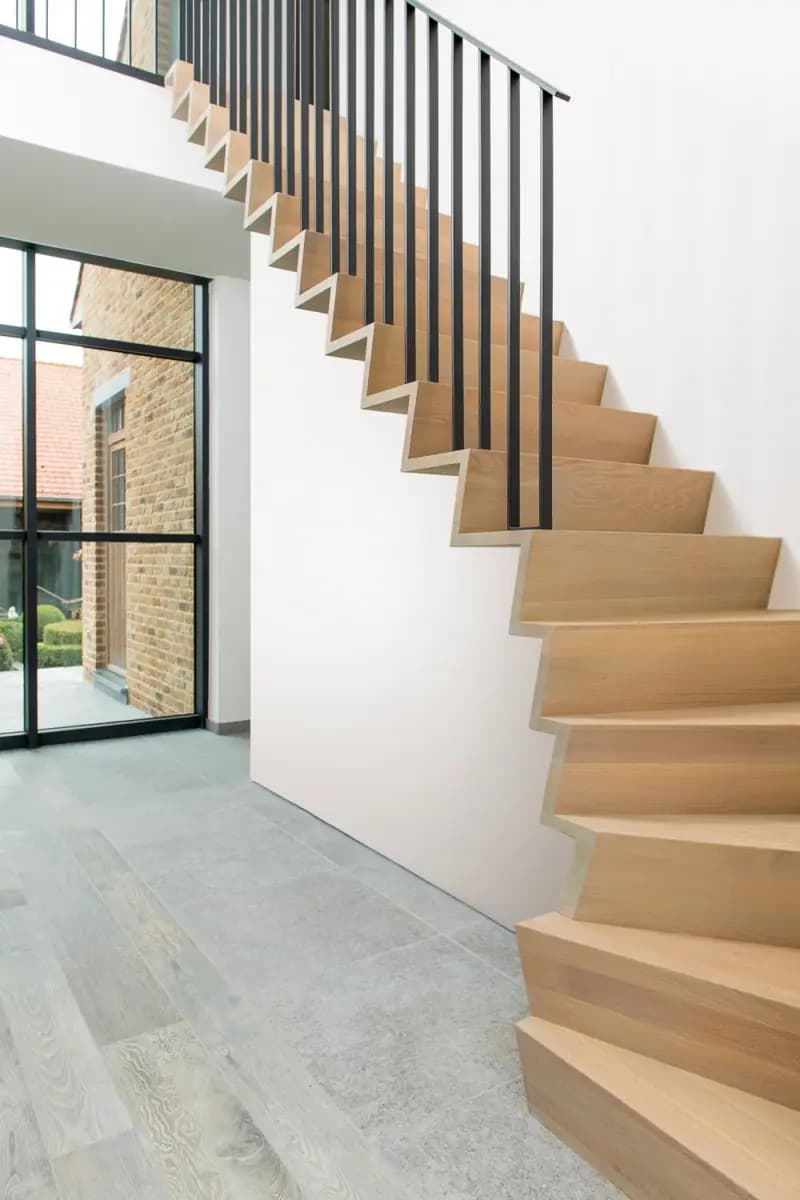 Escalier en bois clair le long d'un mur blanc, garde-corps à fins barreaux noirs, grande baie vitrée à châssis noirs au fond