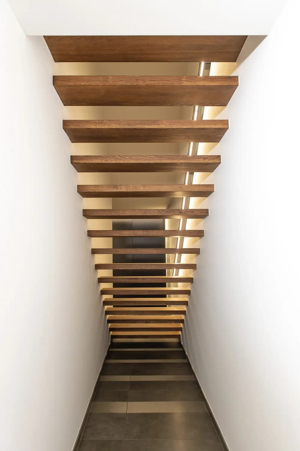 Escalier vu d'en bas entre deux murs blancs rapprochés, marches en bois foncé éclairées par une ligne lumineuse latérale