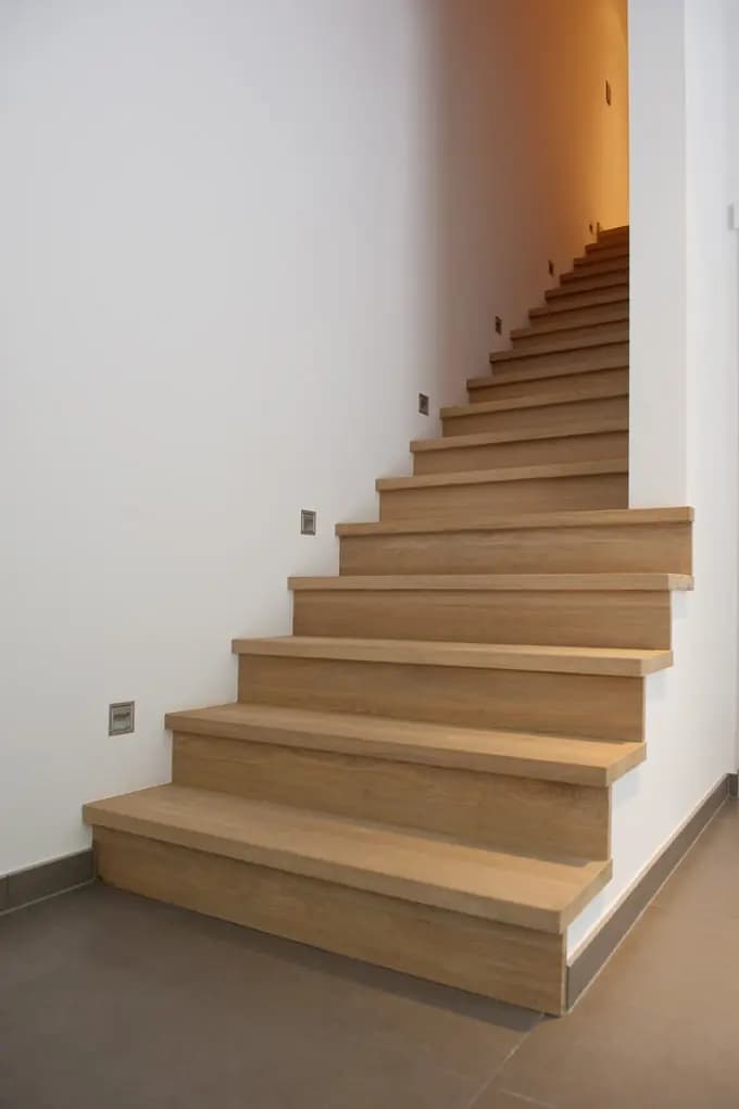Escalier en bois clair aux marches pleines, avec des marches tournantes en bas, longeant un mur blanc
