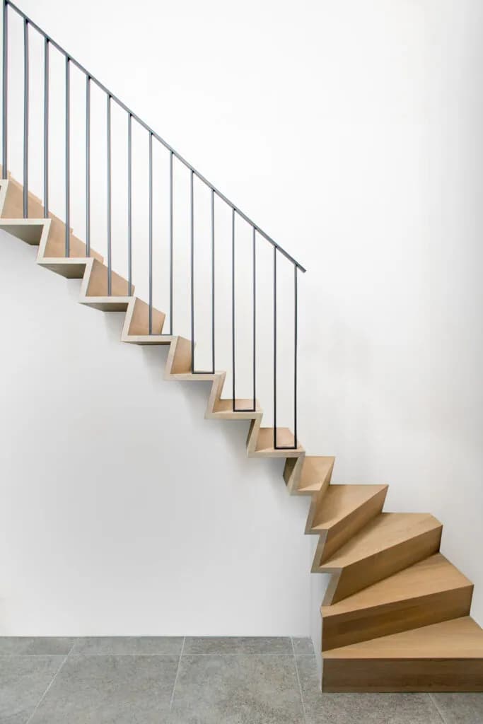 Escalier en bois clair contre un mur blanc, garde-corps fait de fins barreaux métalliques noirs surmontés d'une main courante droite