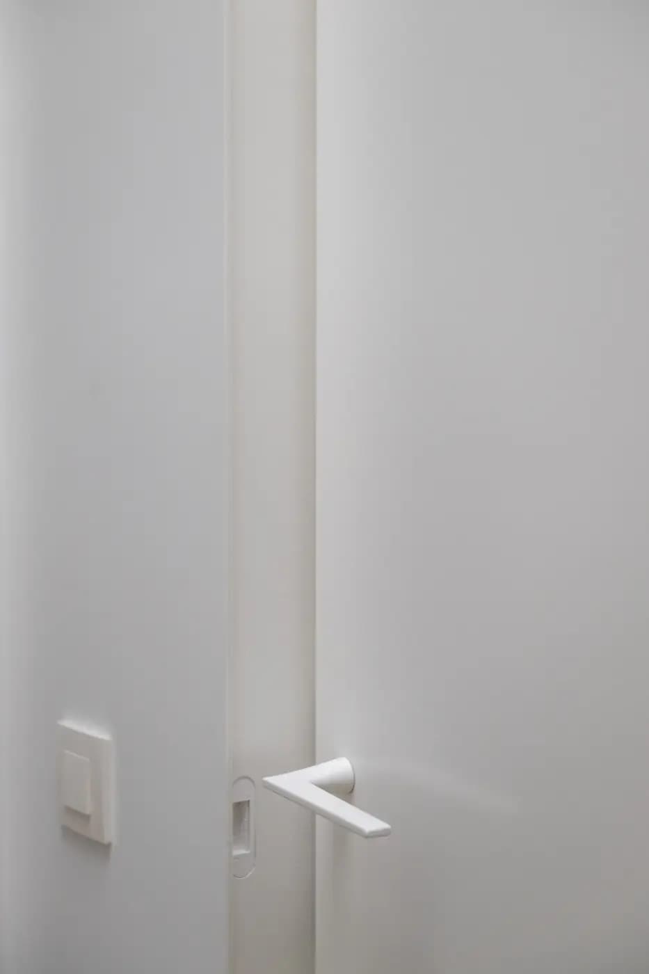 Gros plan sur une porte blanche entrouverte, poignée blanche et interrupteur blanc sur le mur, tout dans la même teinte