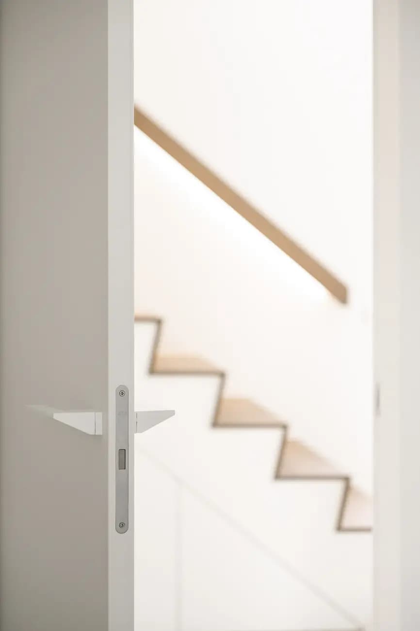 Gros plan sur le bord d'une porte blanche ouverte, escalier en bois clair et main courante flous en arrière-plan