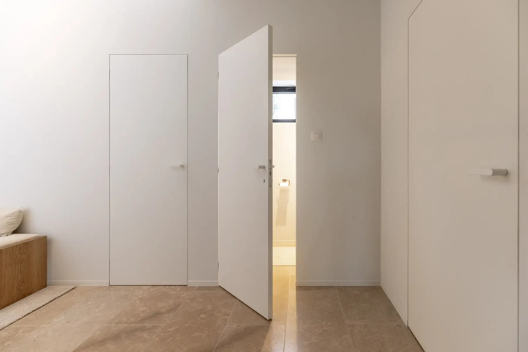 Intérieur clair, portes blanches pleine hauteur prises dans le mur, l'une entrouverte sur une salle d'eau, sol en pierre beige