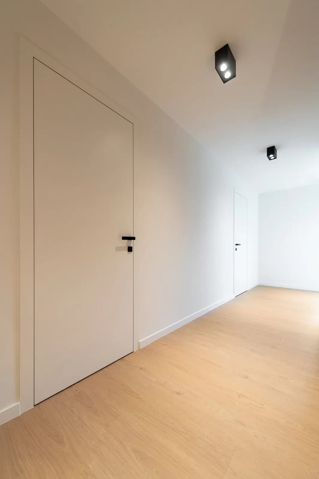 Porte blanche pleine hauteur prise dans un mur clair, poignée noire, couloir clair et sol en bois blond, plafond incliné