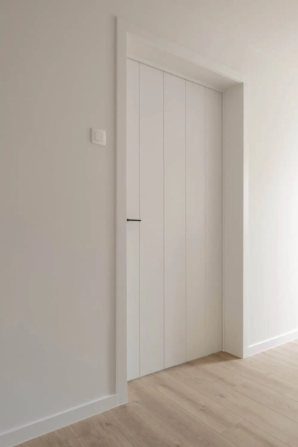 Porte blanche fermée dans un encadrement blanc, panneaux verticaux, interrupteur sur le mur, sol en bois clair