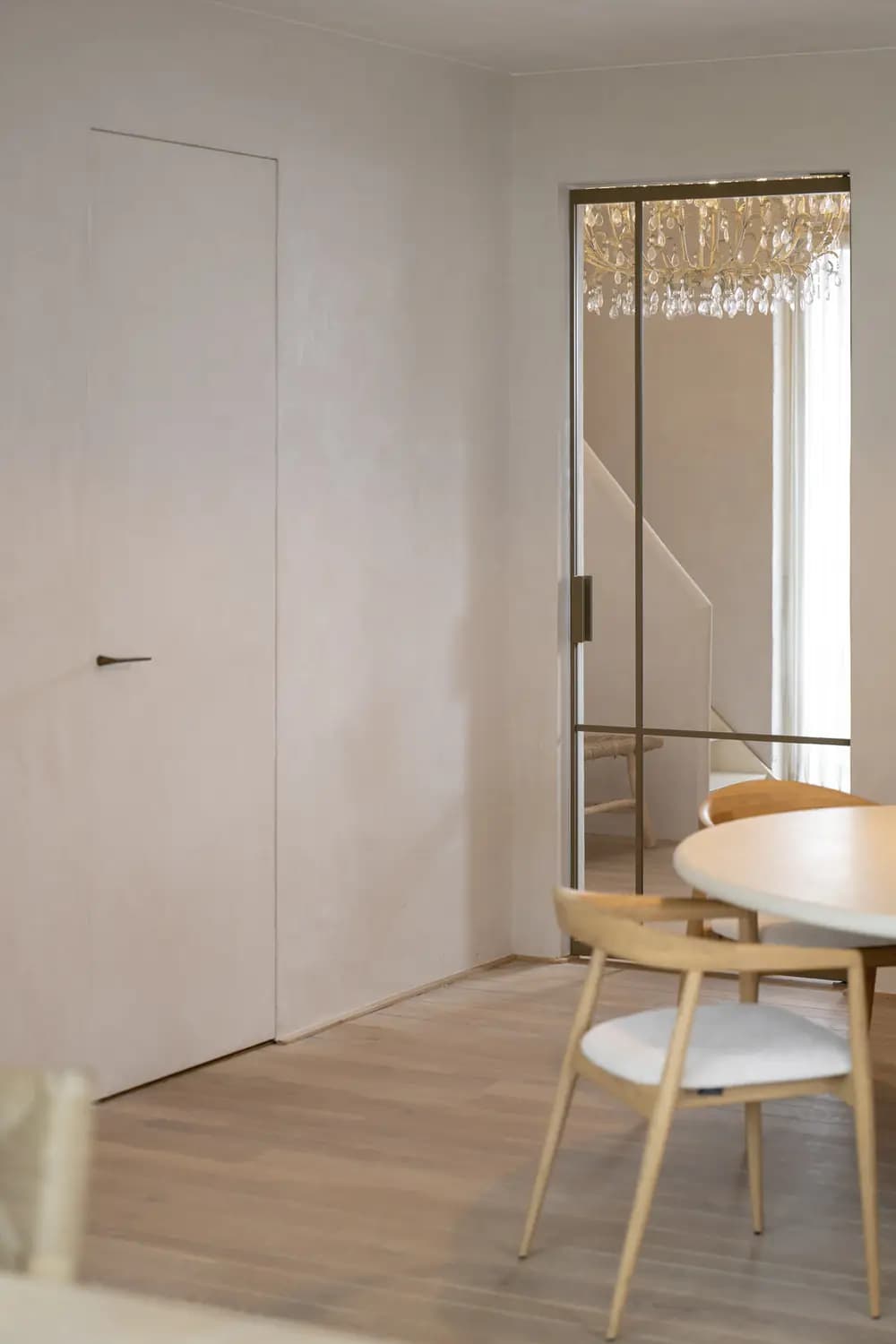 Porte claire pleine hauteur fermée dans un mur de la même teinte, à côté d'une porte vitrée à châssis noir ouvrant sur une salle à manger